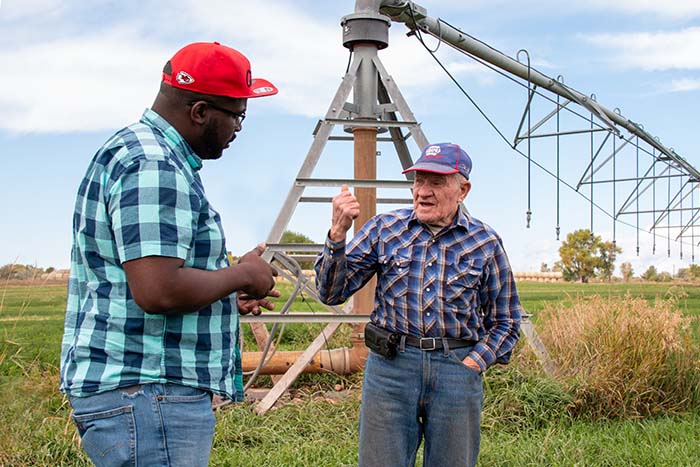 Two men stand in a field and discuss equipment in the background