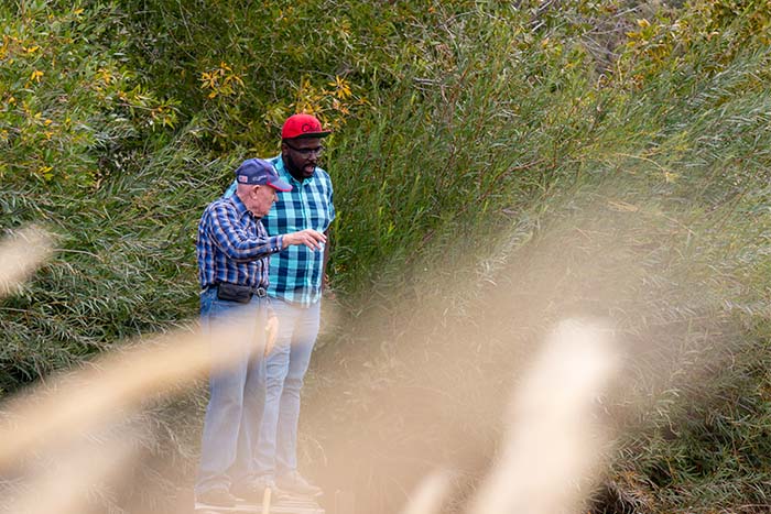 Two men standing on a bridge overlooking a dried lake, with foliage blurred in the foreground