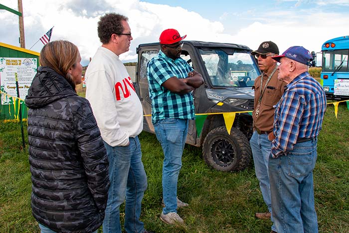 Four men and one woman talk at a fair, with vehicles and flags in the background