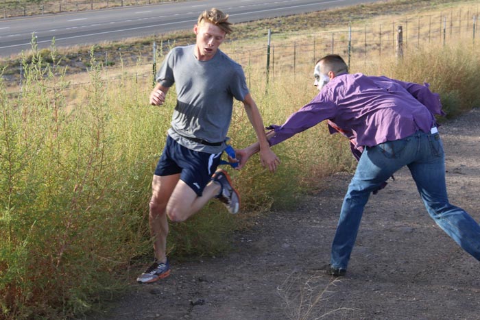 A man in zombie makeup grabs a flag tied around the waist of a runner